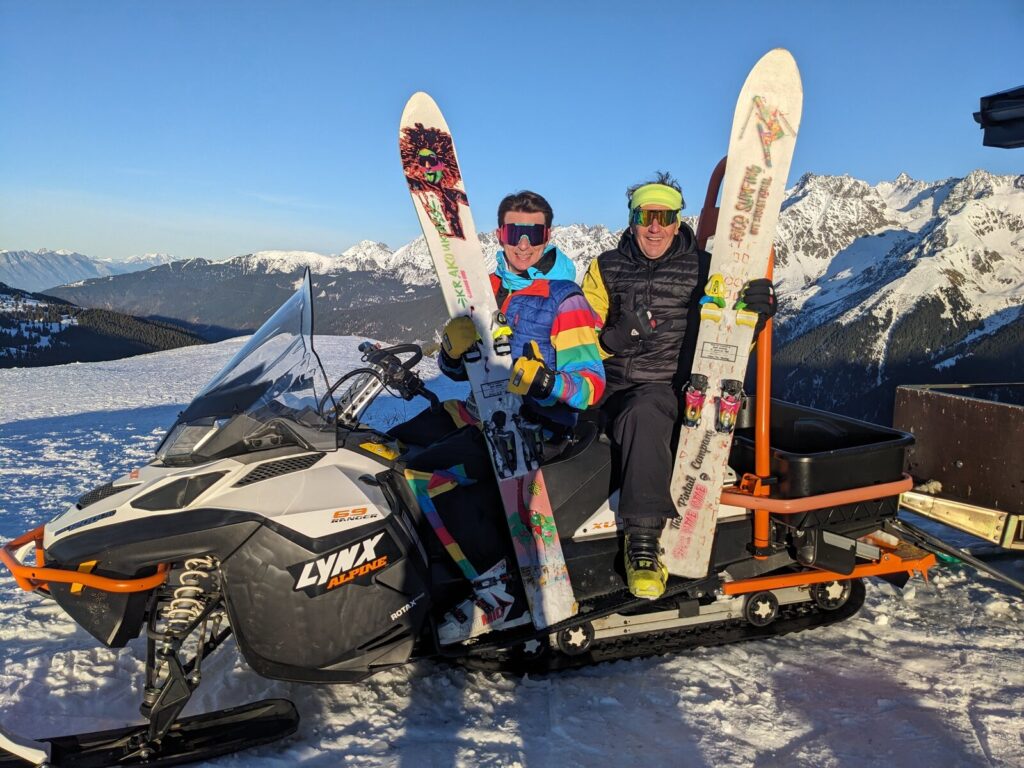 Trois riders posent avec leurs monoskis à côté d’une dameuse, en haut des pistes