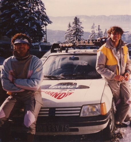 Deux riders en tenue de ski des années 80 appuyés sur une voiture, en bord de piste enneigée