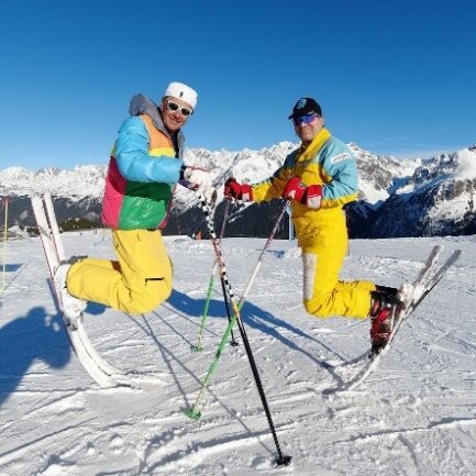 Deux riders en combinaisons fluo, skis aux pieds, posant sur une piste ensoleillée